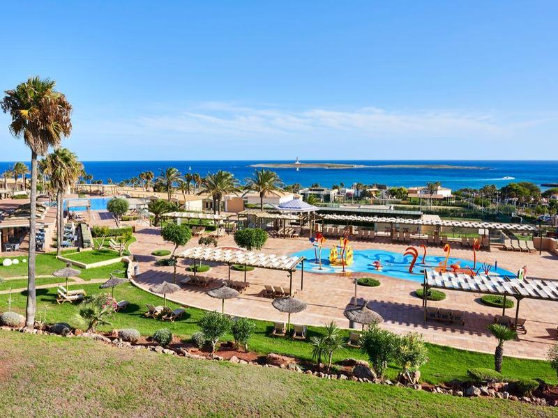 View of a hotel pool with sun loungers, palm trees, and the sea in the background under a blue sky.