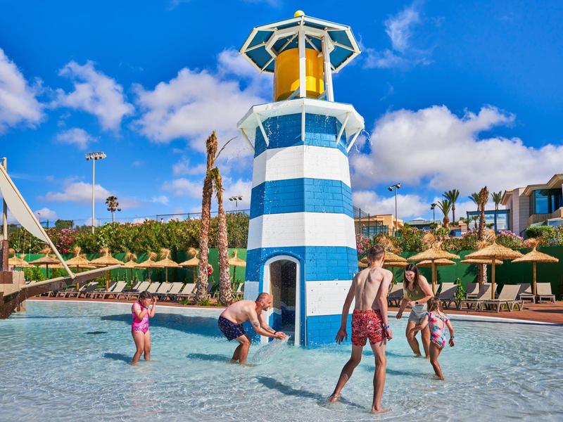 Children playing in a shallow pool with a blue and white lighthouse play structure.
