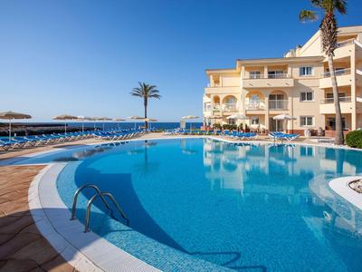 Large hotel pool with sun loungers and sea view under clear sky.