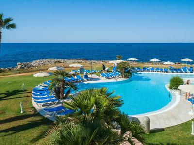 Runder Hotelpool mit Sonnenliegen, Palmen und Meerblick unter klarem Himmel.