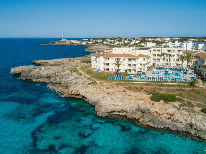 Hotel on rocky coast with pool and turquoise sea under clear sky.