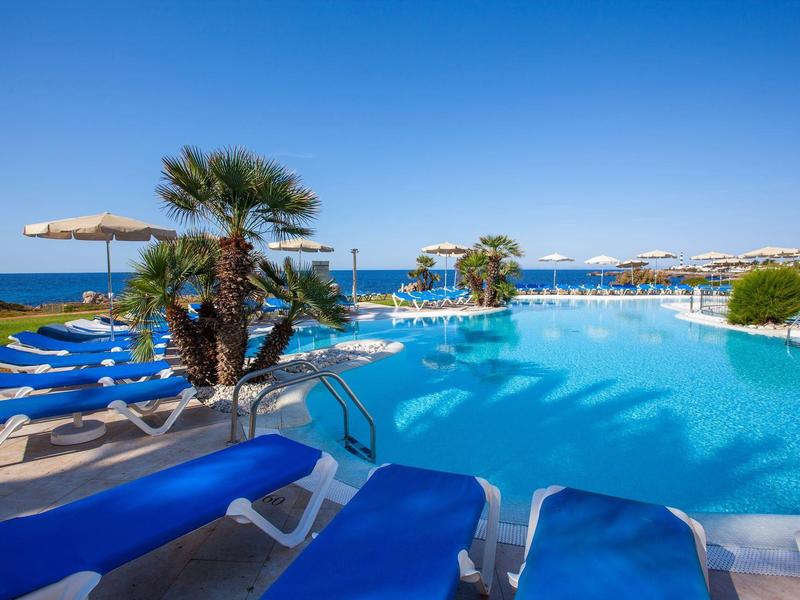 Large outdoor pool with blue lounge chairs and palm trees under clear blue sky.