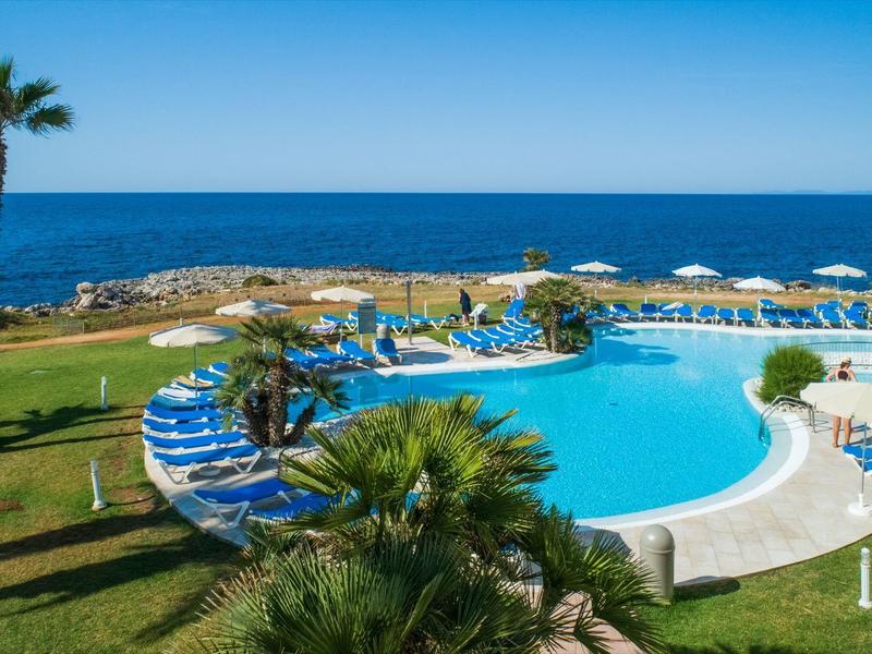 Pool with blue lounge chairs by the sea, surrounded by palm trees and umbrellas