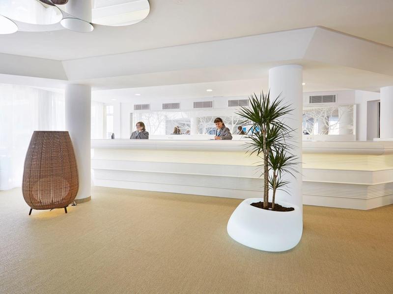 Modern, sunlit hotel lobby with white reception desk and decorative plants.