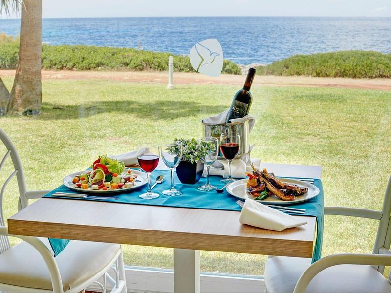 Table with food and wine by a panoramic window overlooking the sea and greenery.
