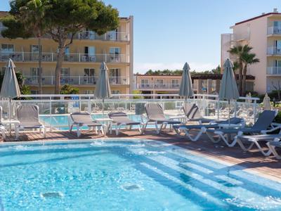 Clear hotel pool with sunbeds and umbrellas in front of yellow buildings under a blue sky.