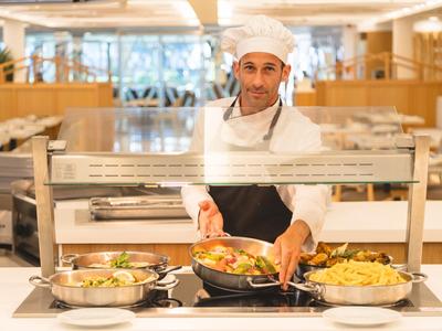 A chef serves freshly prepared dishes from a buffet in a bright dining hall.