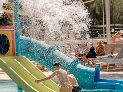 Children play on two slides in a hotel pool with water splashing in the background.