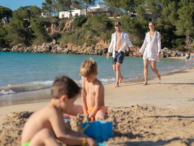 Children play with sand toys on the beach as a couple walks along the water.
