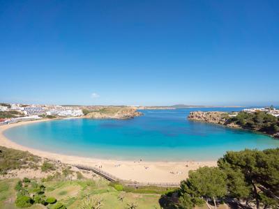 A long sandy beach with clear blue water and a wooded hill in the foreground.