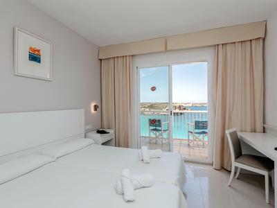 Bright hotel room with two beds, desk, and balcony overlooking the sea.