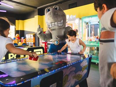 Children playing air hockey with a large mascot in the background.