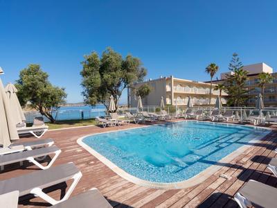 Hotel pool with lounge chairs and umbrellas under blue sky next to olive trees.