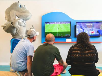 Three people sit in front of two screens watching sports, next to a large shark mascot figure.