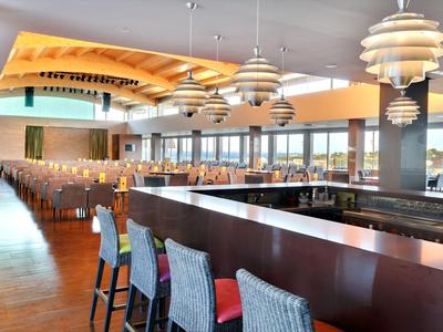 Modernes, helles Restaurant mit langen Holztischen und bunten Stühlen unter weißen Deckengestellen.