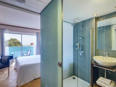 Nowoczesny pokój hotelowy z łóżkiem, częścią wypoczynkową, szklaną kabiną prysznicową oraz widokiem na taras i morze.