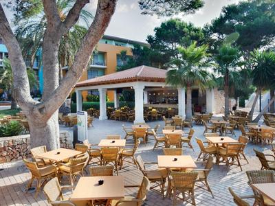 Zewnętrzny obszar hotelu z wieloma drewnianymi stołami i krzesłami pod dużymi drzewami.