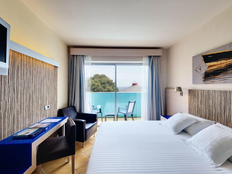 Nowoczesny pokój hotelowy z podwójnym łóżkiem, telewizorem, biurkiem i balkonem z widokiem na morze.