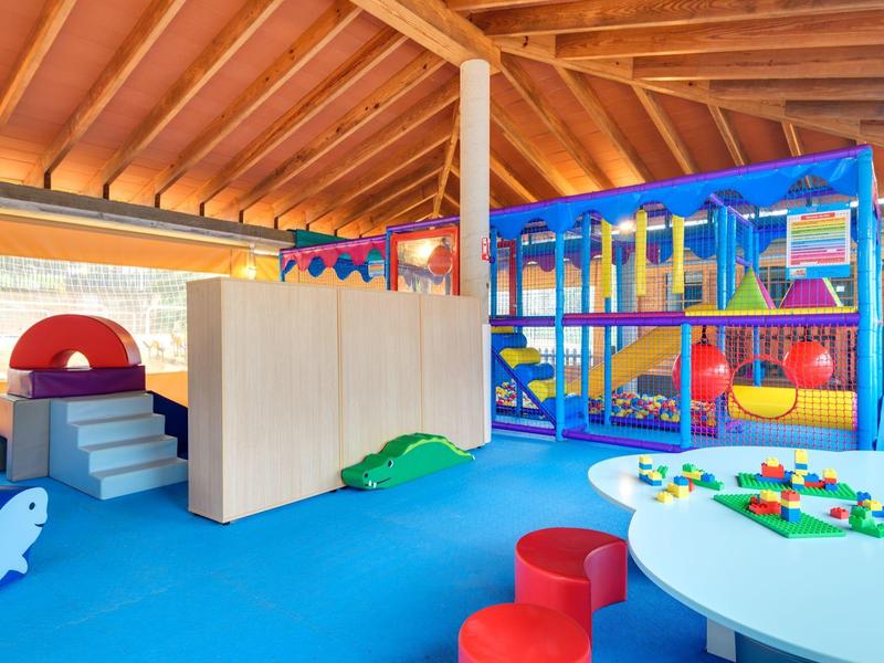 Kolorowy kryty plac zabaw z drabinką do wspinaczki, zjeżdżalnią i stołem do zabawy pod drewnianym dachem.