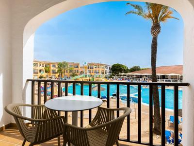 Balkon mit Tisch und Stühlen mit Blick auf den Pool und Hotelgebäude bei sonnigem Wetter.