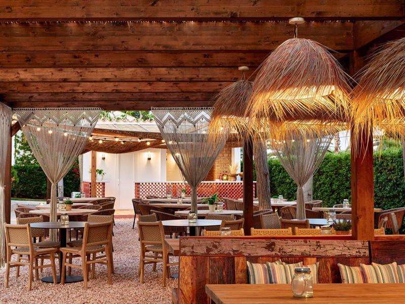 Zastřešená venkovní oblast s dřevěným nábytkem a osvětlením z slaměných deštníků v hotelové restauraci