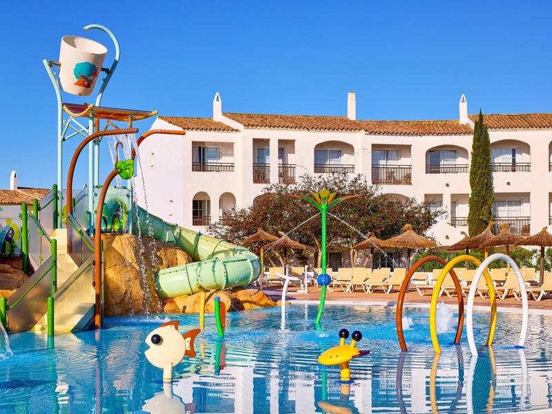 Barevné vodní hřiště s klouzačkami a malými figurkami před hotelem za jasné oblohy.