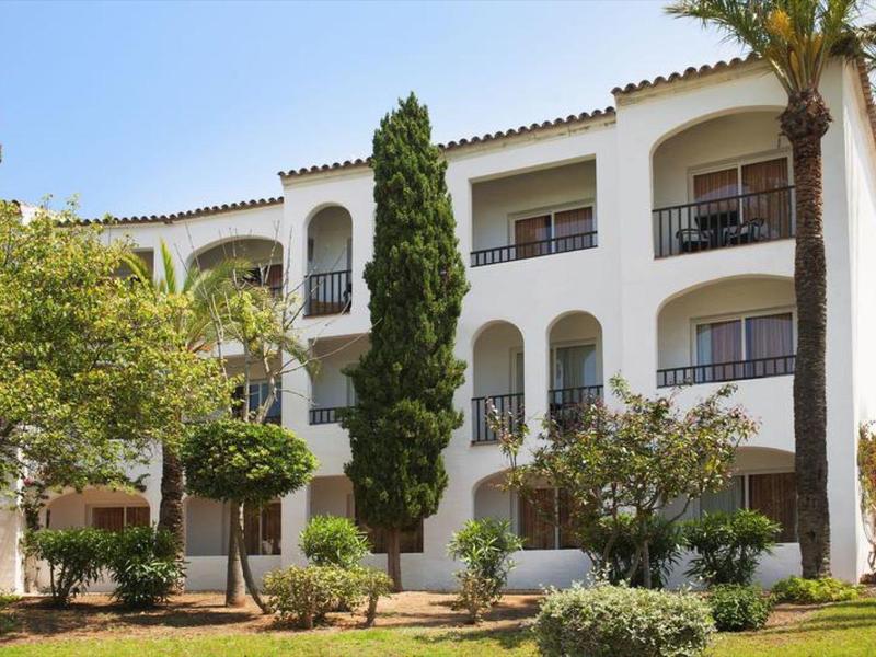 Bílá hotelová budova s oblouky, balkony a středomořskými stromy na dvoře.