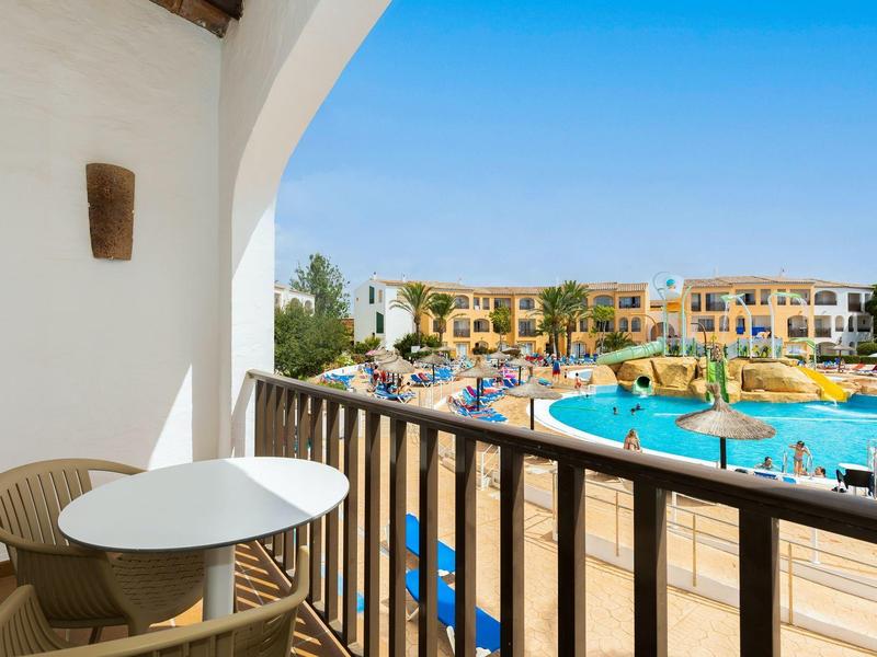 Balkón se stolem a židlemi s výhledem na velký bazén a žluté prázdninové apartmány pod jasnou oblohou.