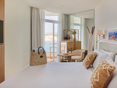 Lichte hotelkamer met comfortabel bed, fauteuils en groot raam met zeezicht.