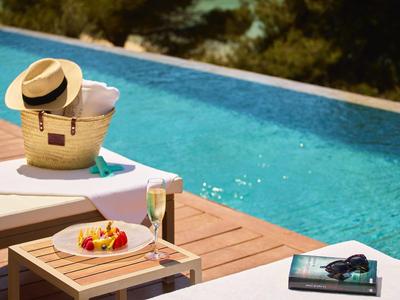 Ligstoel bij infinity pool met strohoed, drankje en fruitbord op houten terras.