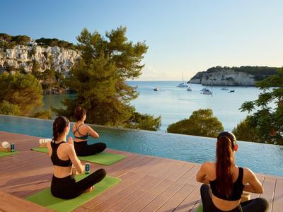 Drei Frauen praktizieren Yoga auf einer Terrasse mit Blick auf das Meer und Boote bei Sonnenuntergang.