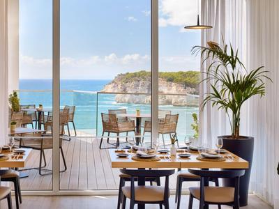 Modernes Restaurant mit Meerblick und Terrasse mit Tischen und Stühlen, Pflanzen und Tageslicht.