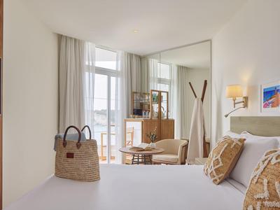 Helles Hotelzimmer mit Doppelbett, dekorativen Kissen und Blick auf Balkon mit Stühlen.