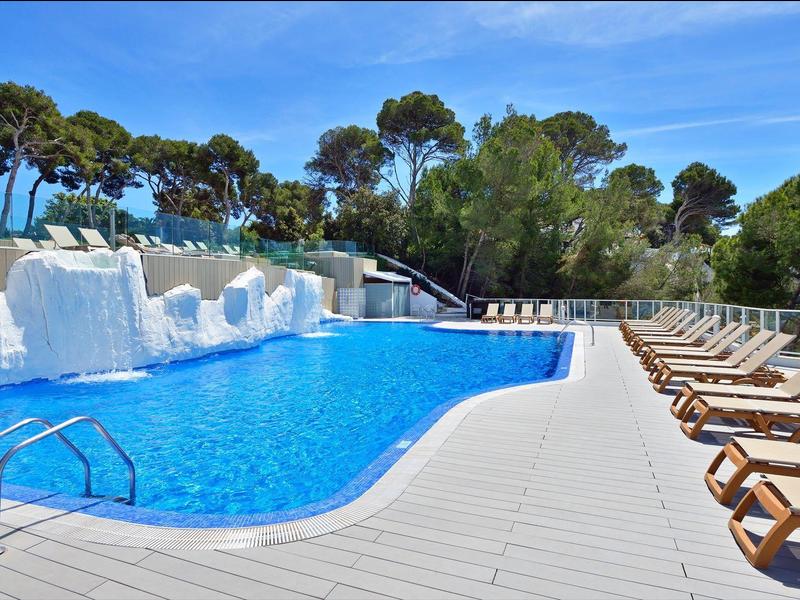Modern hotelzwembad met ligstoelen omgeven door bomen onder een blauwe lucht.