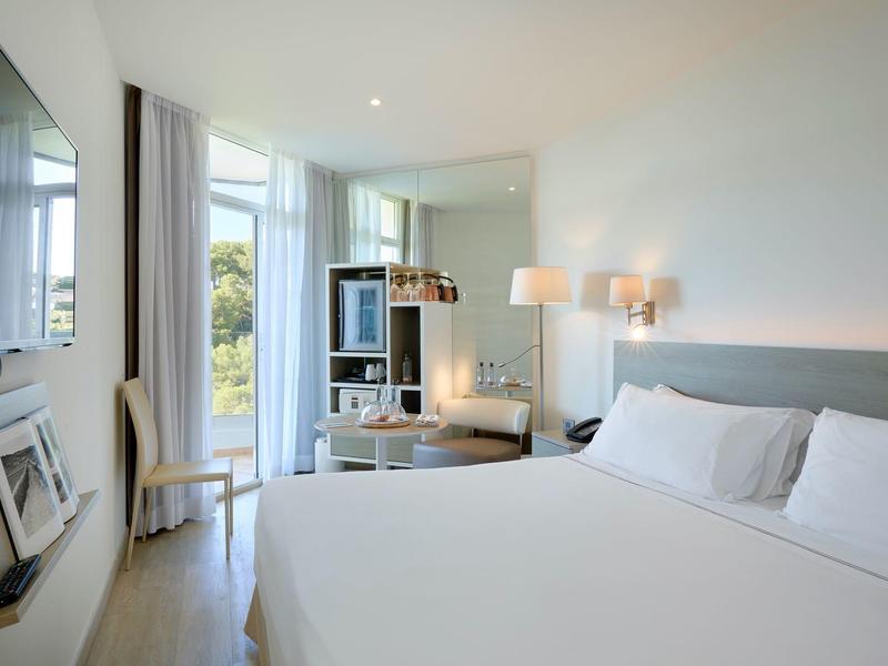 Modern hotelkamer met groot bed, stoel, tv en balkon met uitzicht naar buiten.