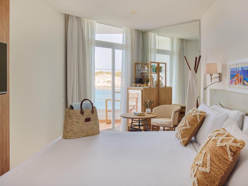 Lichte hotelkamer met comfortabel bed, fauteuils en groot raam met zeezicht.