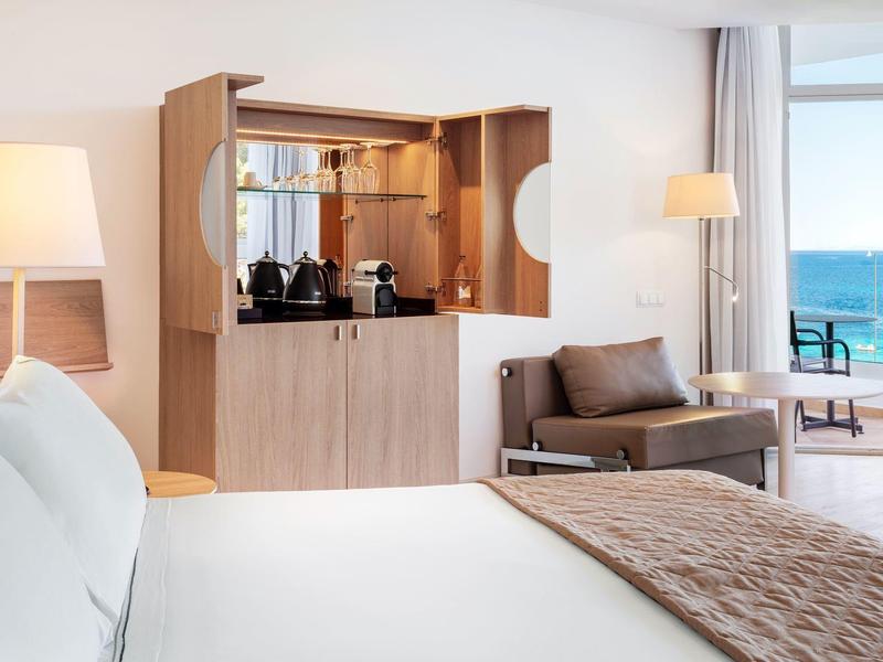 Lichte hotelkamer met groot bed, modern houten barkastje en zeezicht vanaf het balkon.