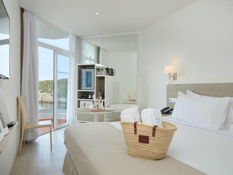 Lichte hotelkamer met groot bed, mandje op het beddengoed en panoramische ramen met uitzicht op het water.