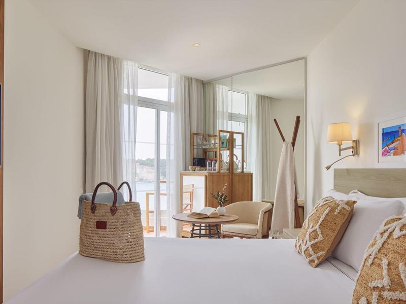 Lichte hotelkamer met tweepersoonsbed, decoratieve kussens en balkon met stoelen.