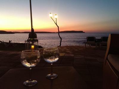 Dos copas de vino en una mesa con puesta de sol sobre el mar y la playa al fondo.