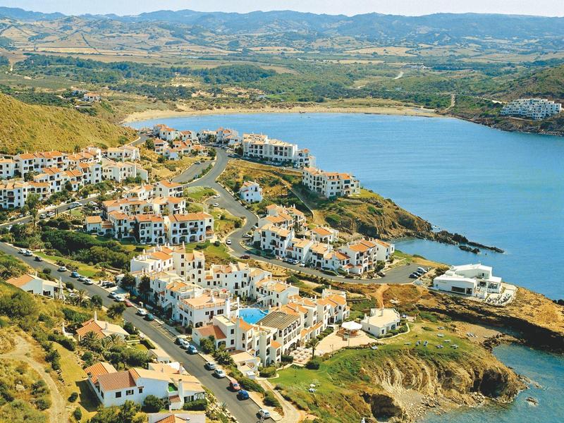 Resort costero con varios edificios con vistas al mar azul en un paisaje montañoso.