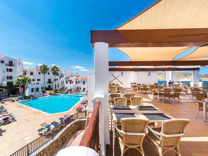 Terraza del hotel con zona de estar con vistas a la piscina y edificios blancos bajo un cielo azul.