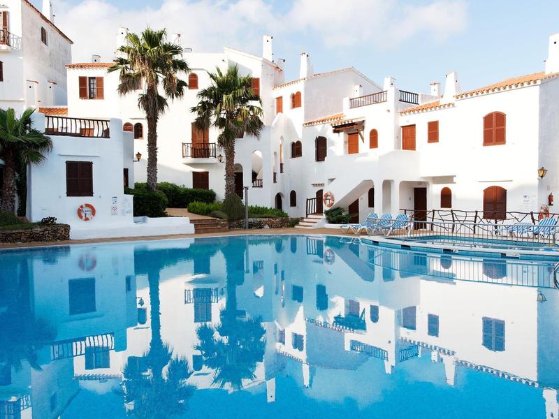 Hotel blanco con palmeras alrededor de la piscina azul bajo un cielo nublado.