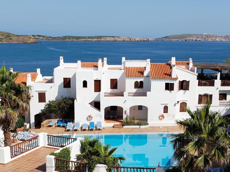 Hotel blanco con techos rojos, piscina y palmeras frente al mar azul y cielo despejado.