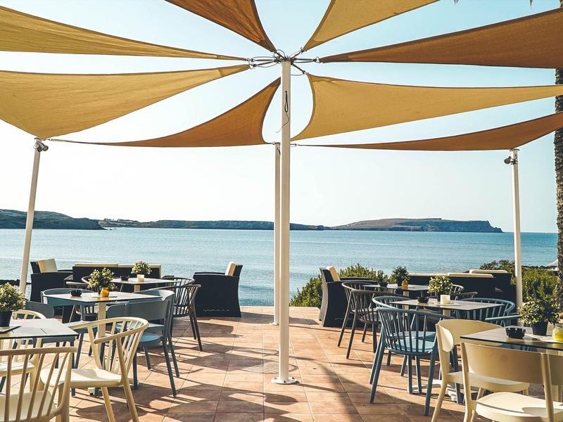 Terraza abierta con mesas y sillas con vista al mar bajo velas de sol.
