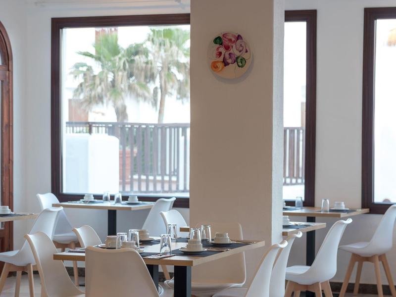 Restaurante luminoso con sillas blancas, mesas y grandes ventanales con vista a palmeras.