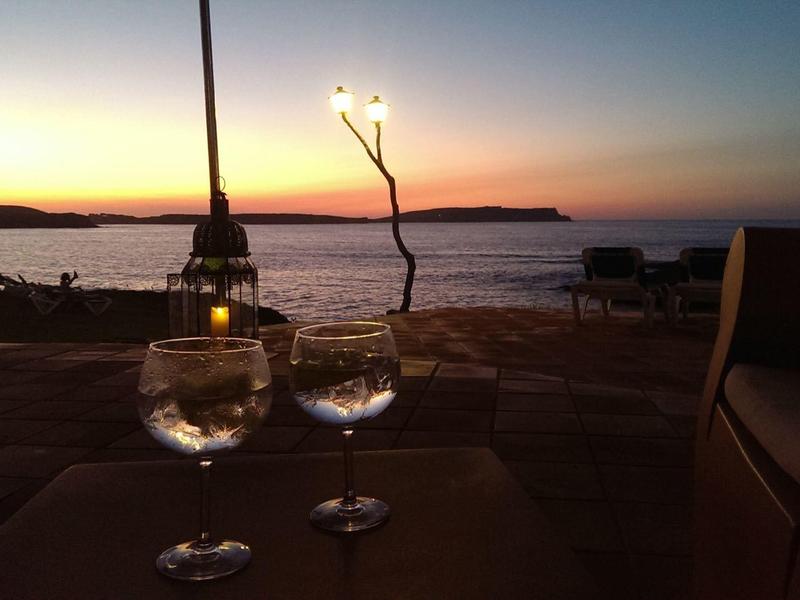 Dos copas de vino en una mesa con puesta de sol sobre el mar y la playa al fondo.