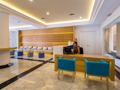 Helle Hotel-Lobby mit Rezeption, blauen Sesseln und gepolsterten Sitzbänken an der Wand.