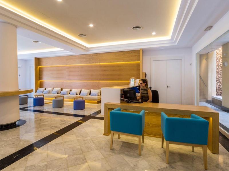 Helle Hotel-Lobby mit Rezeption, blauen Sesseln und gepolsterten Sitzbänken an der Wand.