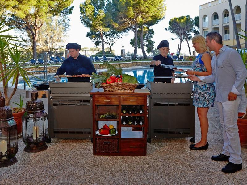Zwei Köche grillen im Außenbereich, während ein Paar ihnen beim Hotelrestaurant zusieht.